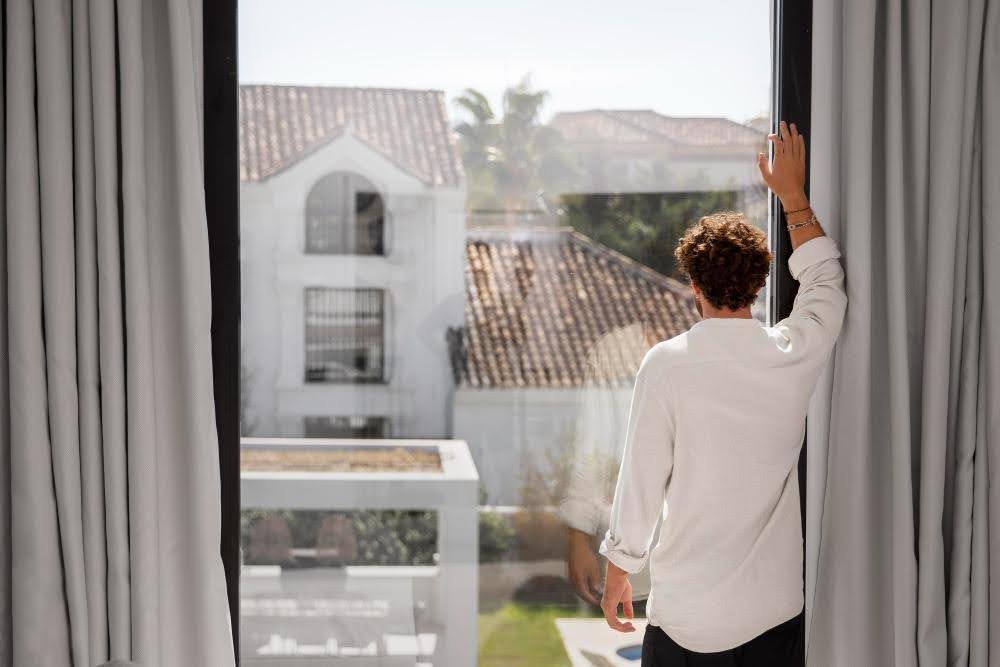 A man facing a large window with drapes pulled aside.