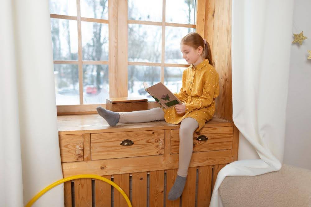 A young girl sitting by a window with heavy drapes - one of the best window treatments for winter.