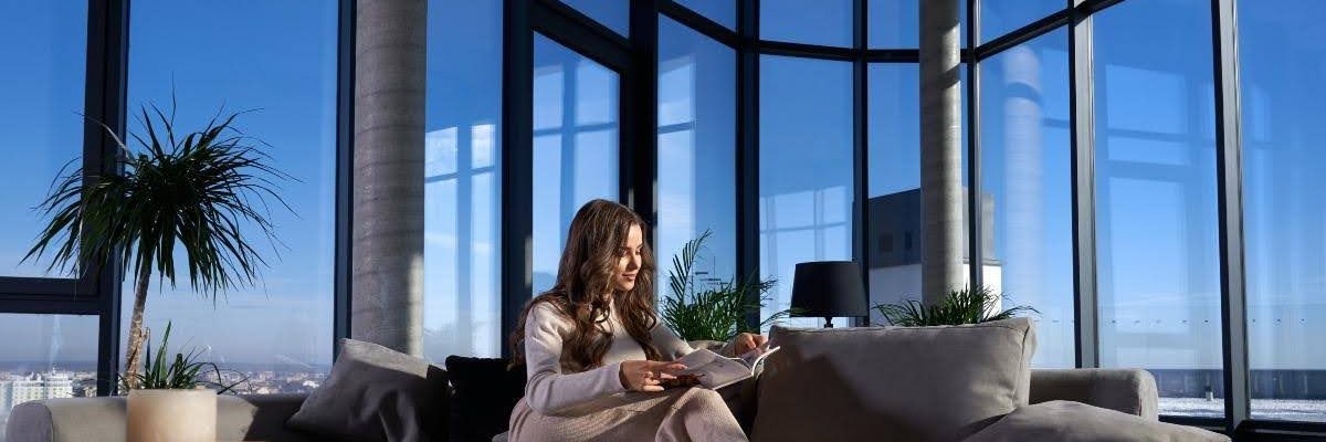 Woman reading a book on a sofa in front of big windows.