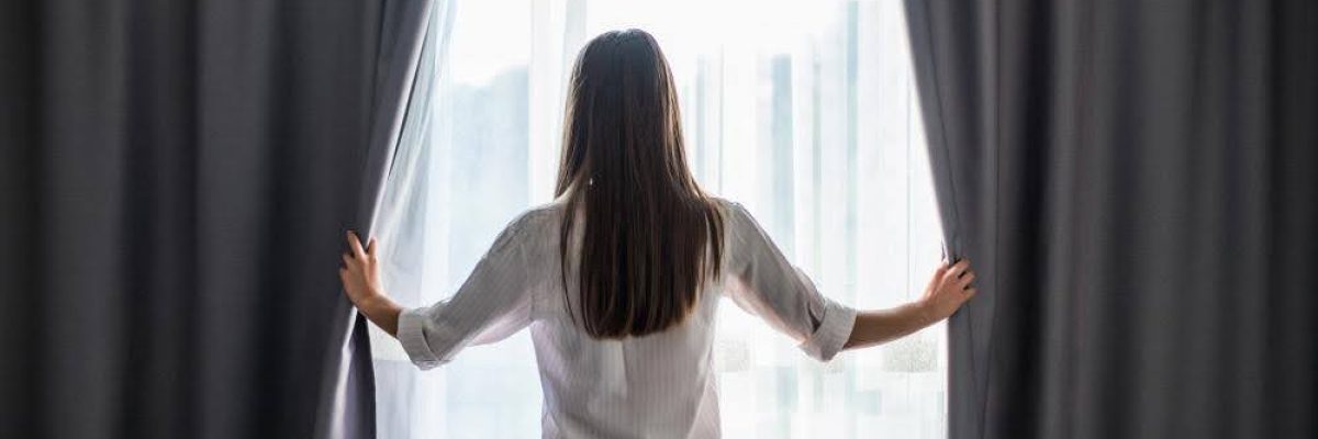 A woman pulling back curtains to let light into the room.