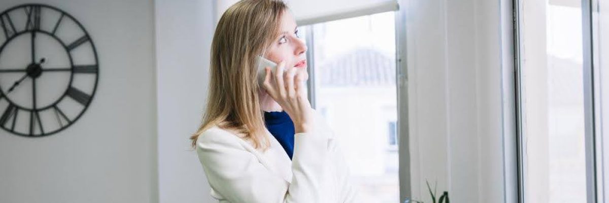 A woman talking on the phone while standing by a window with partially lowered shades.