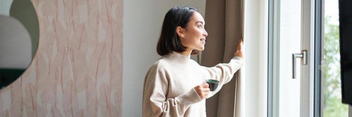 A woman standing next to a window with layered shades.