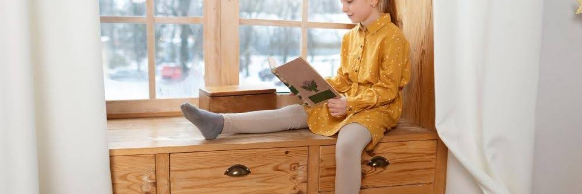 A young girl sitting by a window with heavy drapes - one of the best window treatments for winter.